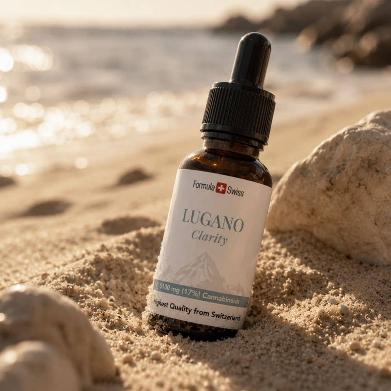 Formula Swiss Lugano Clarity CBD oil bottle displayed in beach sand beside rocks and sunlit water.